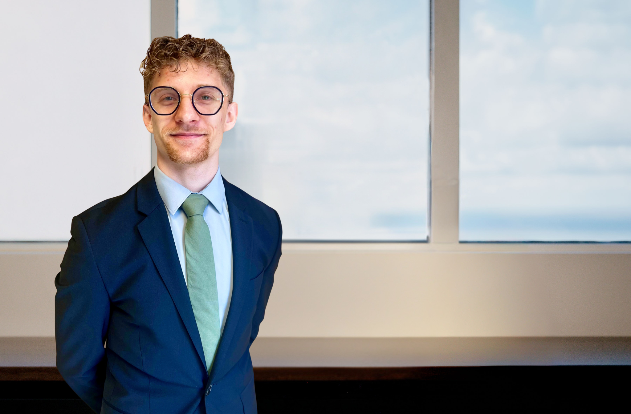 Young man with curly red hair and round glasses wearing a navy suit, light blue shirt, and green tie, smiling before a window
