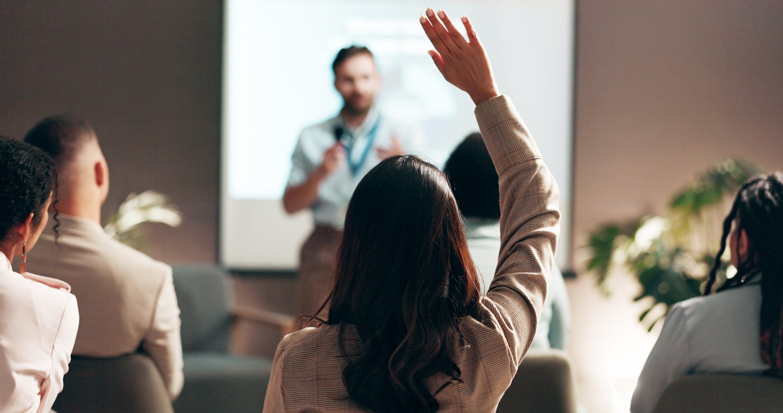 Woman with long dark hair raising her hand at a business conference while a male speaker holds a microphone near a project...