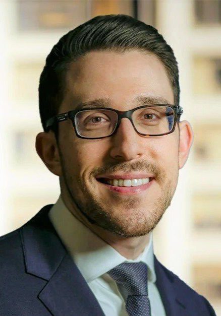 Smiling man with dark hair, glasses, and stubble wearing a navy suit and gray tie in front of a blurred city building window