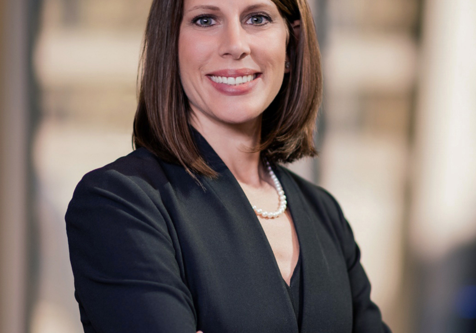 Woman with brown hair in black blazer and pearl necklace smiling with arms crossed in a professional headshot