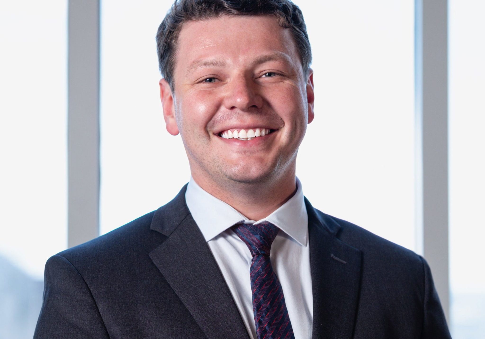 Smiling young man in dark suit, white dress shirt, and red patterned tie, posed for professional headshot near bright offi...