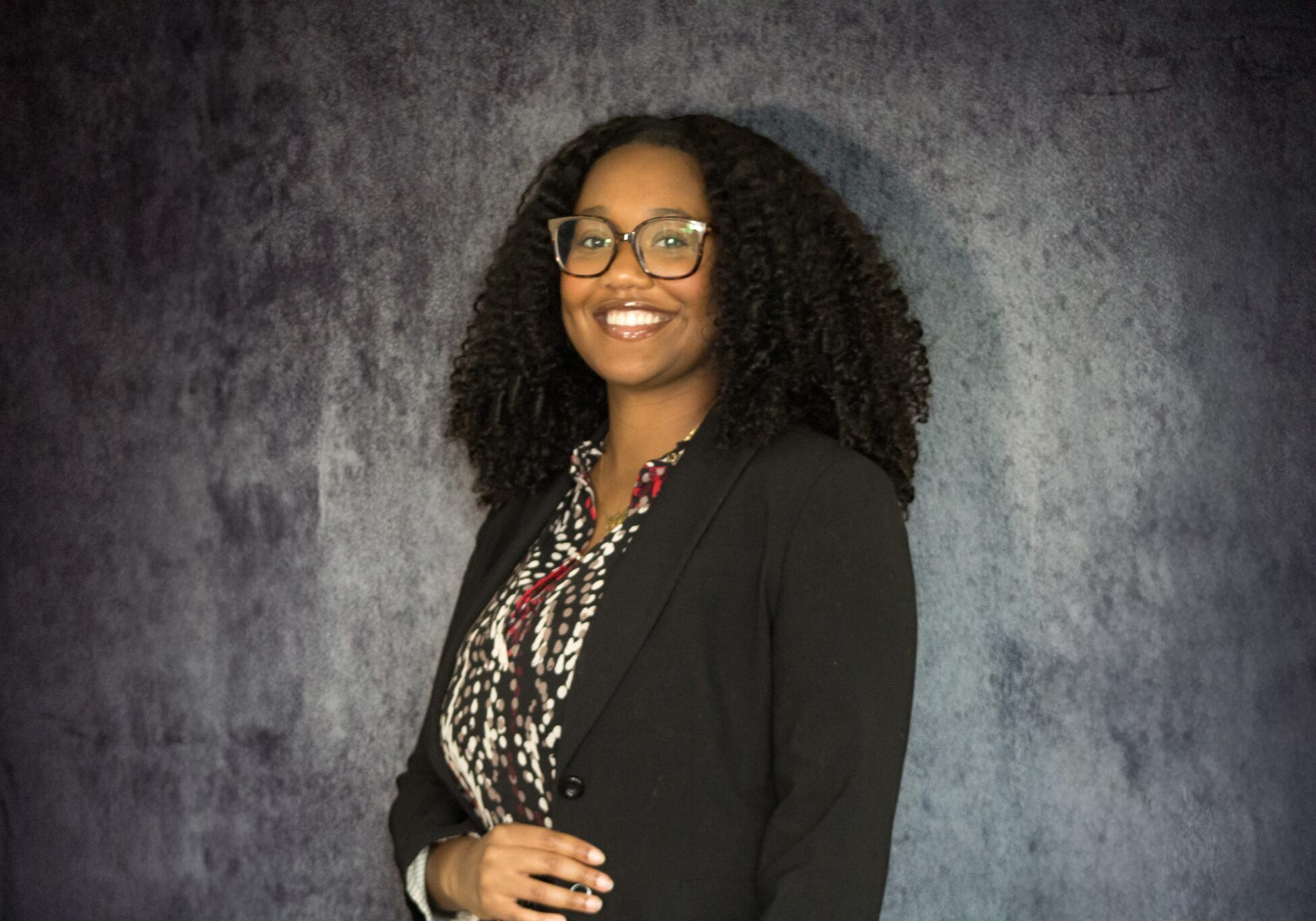 Smiling young woman with curly hair and glasses wearing a black blazer over a patterned blouse, posed against a dark gray ...