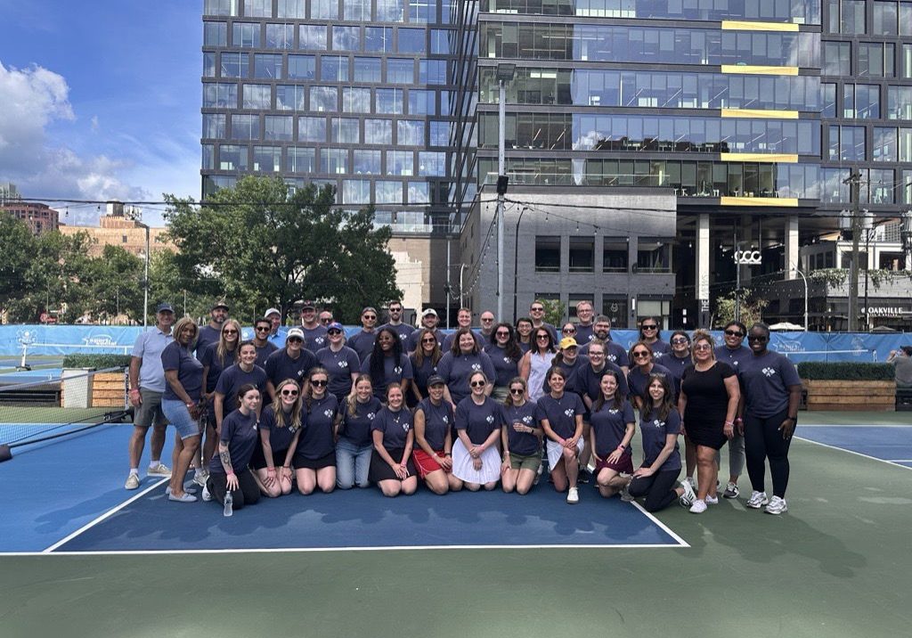 Large group of people in matching navy blue t-shirts posing on an outdoor pickleball court with a modern glass skyscraper ...