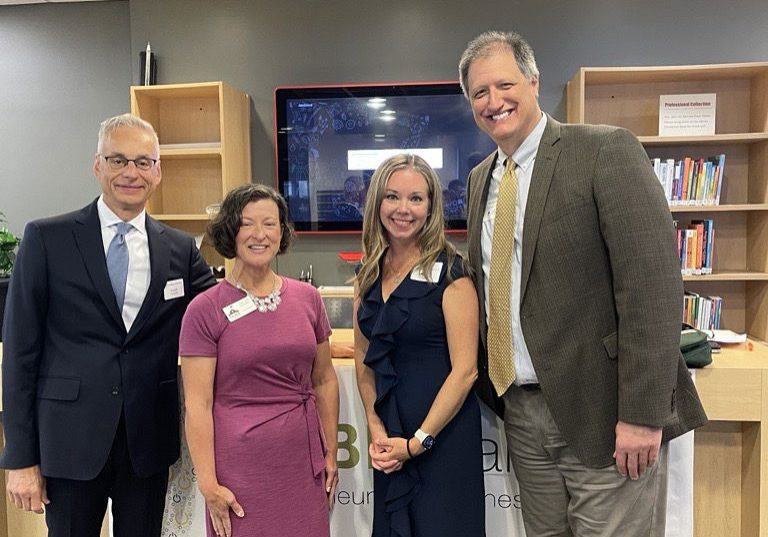 Four professionals smiling together in a modern library or learning center with wooden shelving and a mounted TV screen be...