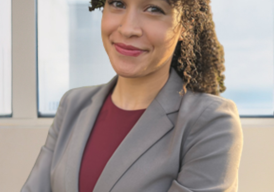 Woman with curly hair smiling with arms crossed, wearing a gray blazer and burgundy top, standing near an office window