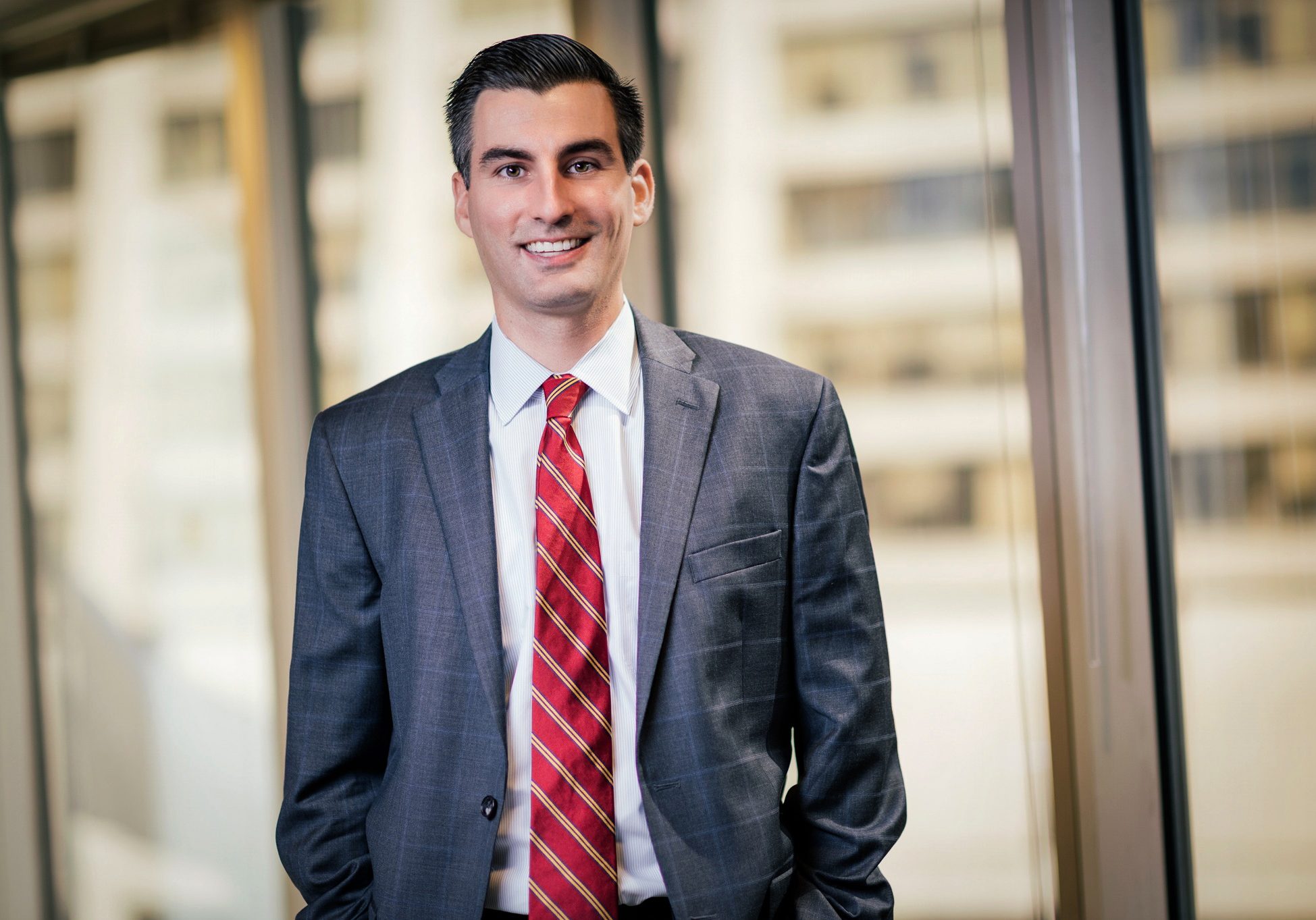 Young man in gray suit and red striped tie smiling confidently near floor-to-ceiling office building windows