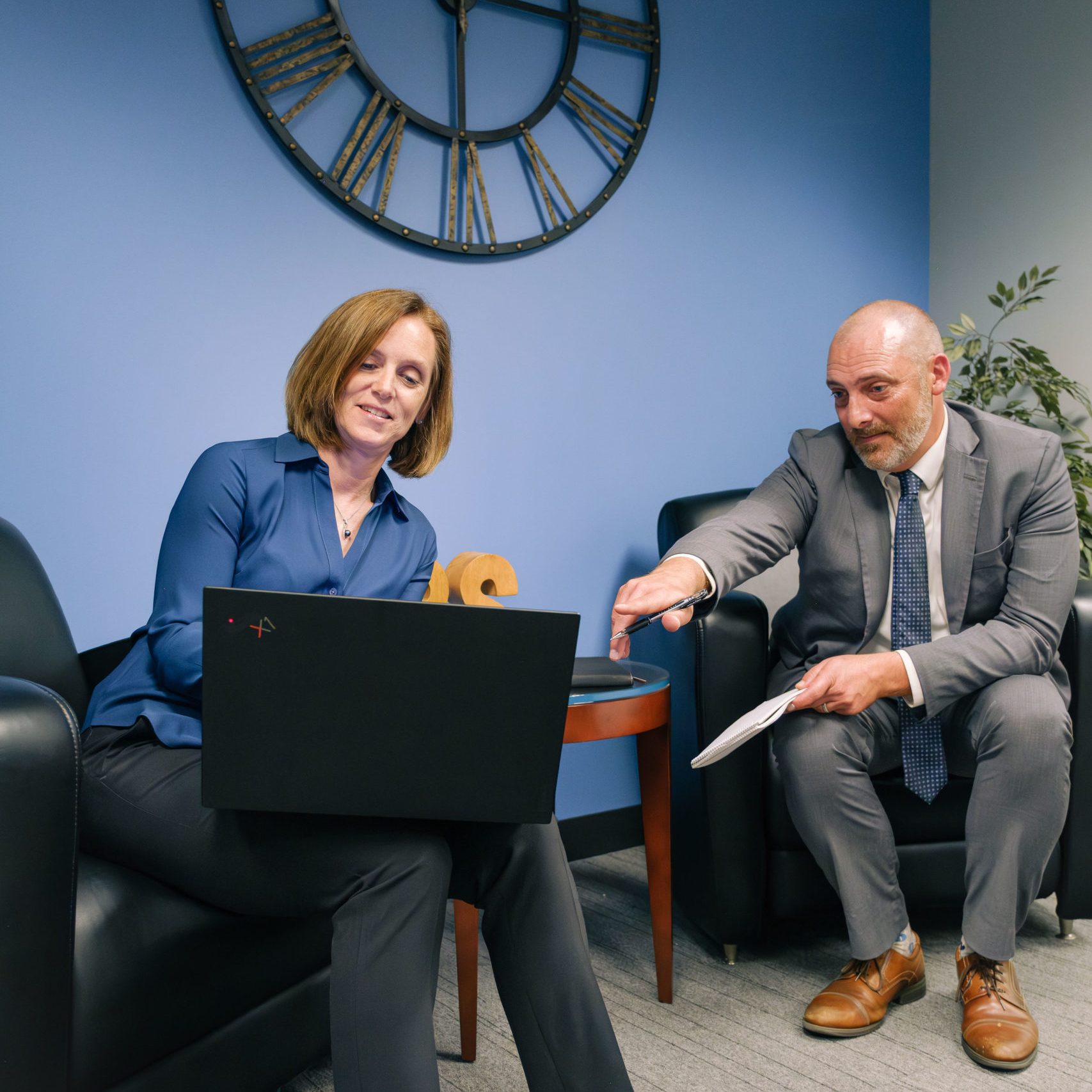 Woman with laptop and man in gray suit reviewing documents in a blue office with a large Roman numeral wall clock