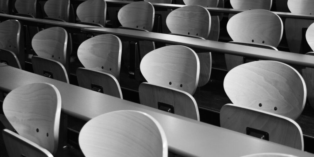 Rows of empty wooden chairs with curved backs in a lecture hall or auditorium, photographed in black and white