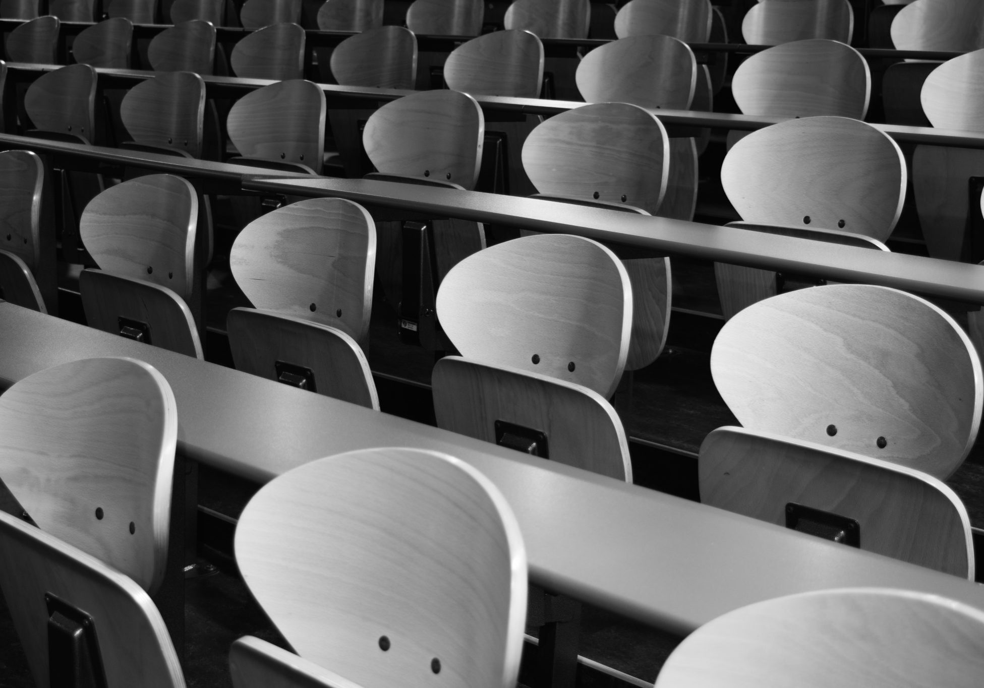 Rows of empty wooden chairs with attached desks in a lecture hall, photographed in black and white