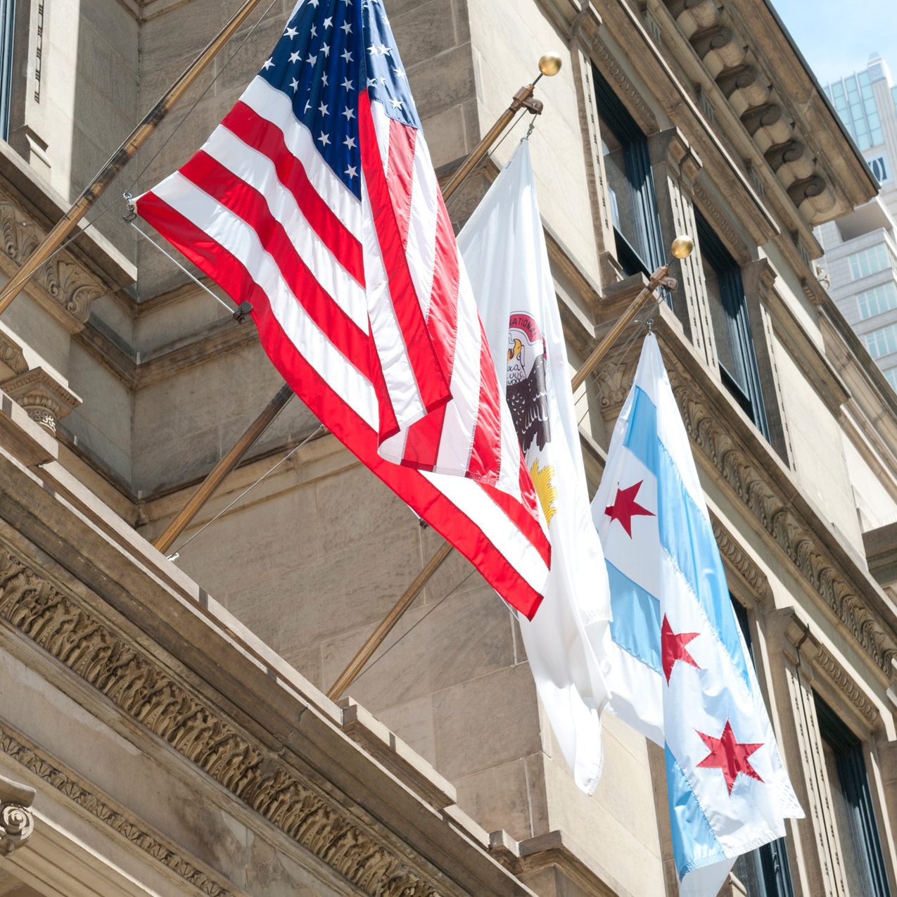 American flag, Illinois state flag, and Chicago city flag displayed on a ornate stone government building with skyscrapers...