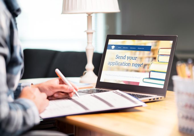 Person writing on documents at a desk beside a laptop displaying a university website with "Send your application now!" on...