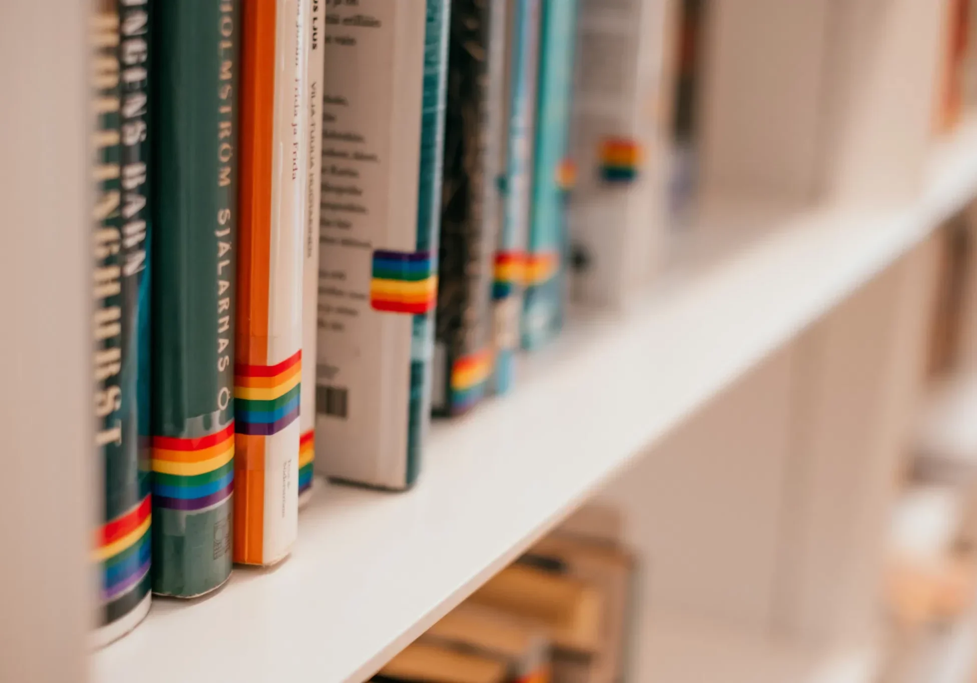 Library shelf with books marked by small rainbow Pride flag stickers on their spines, indicating LGBTQ+ themed or inclusiv...