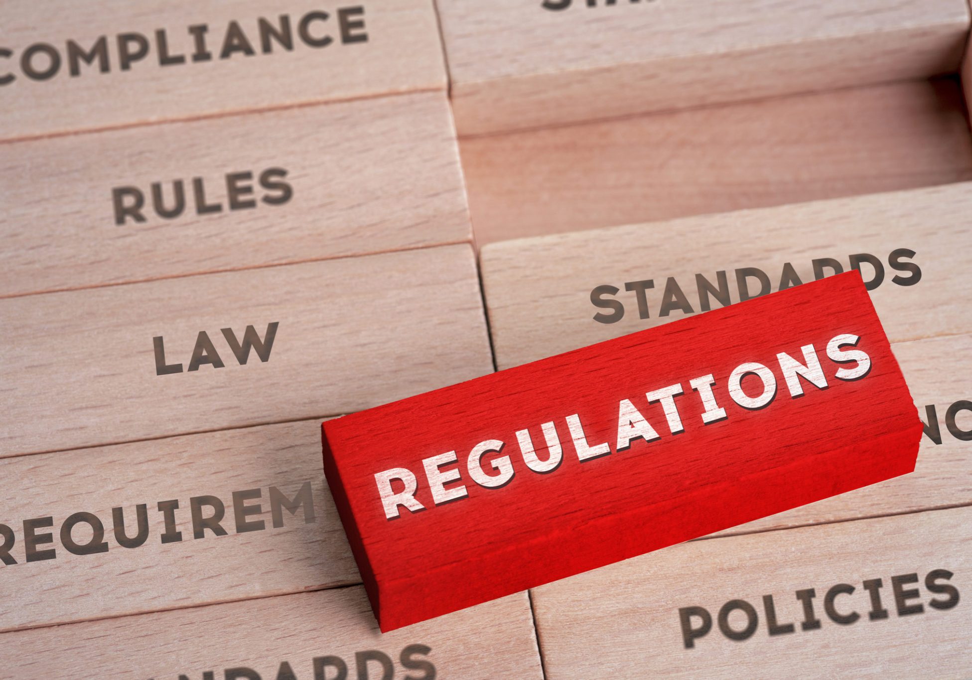 Red wooden block labeled REGULATIONS lying atop beige blocks stamped with Compliance, Rules, Law, Standards, Policies, and...