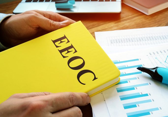 Person holding a yellow EEOC book over a desk with bar chart reports, a blue marker, and a laptop in the background.