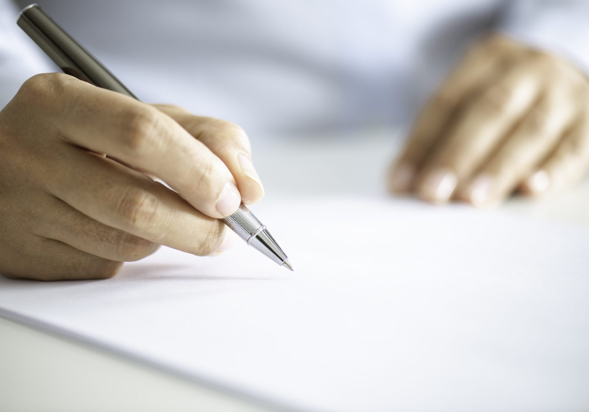 Person in white shirt writing on blank white paper with a silver ballpoint pen, close-up view of hands on desk
