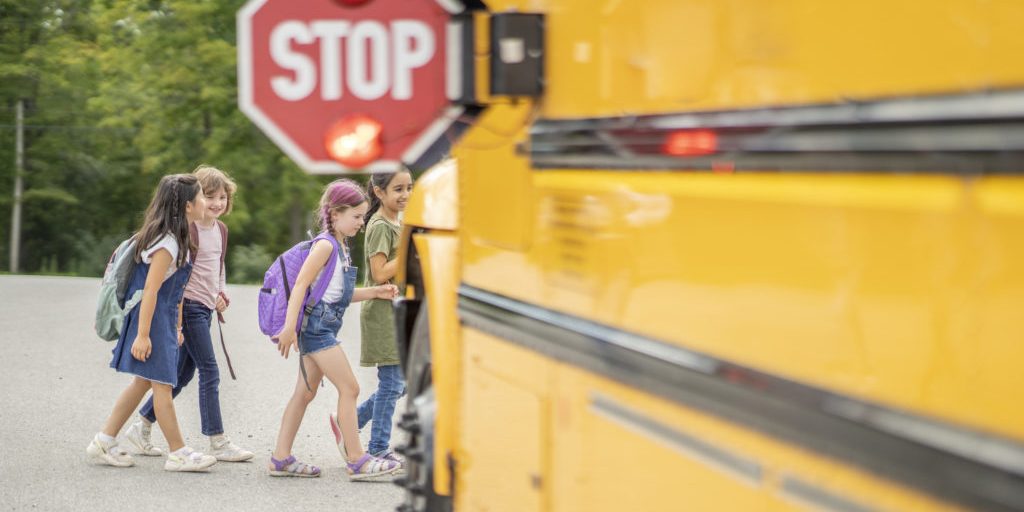 Four young girls with backpacks boarding a yellow school bus with extended STOP sign and flashing red lights on a rural road.