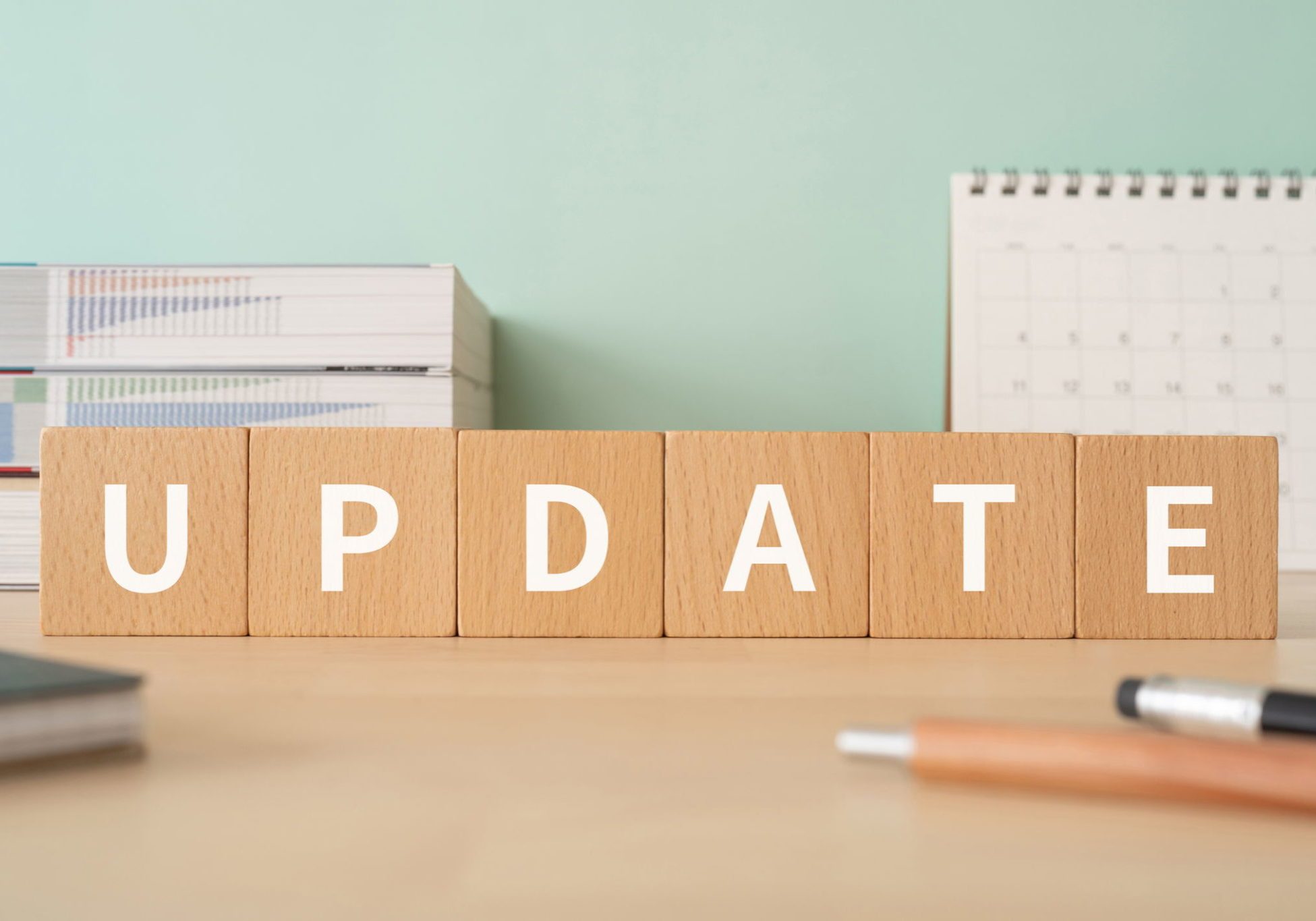 Wooden letter blocks spelling UPDATE on a desk with stacked papers, a calendar, and pens in the background