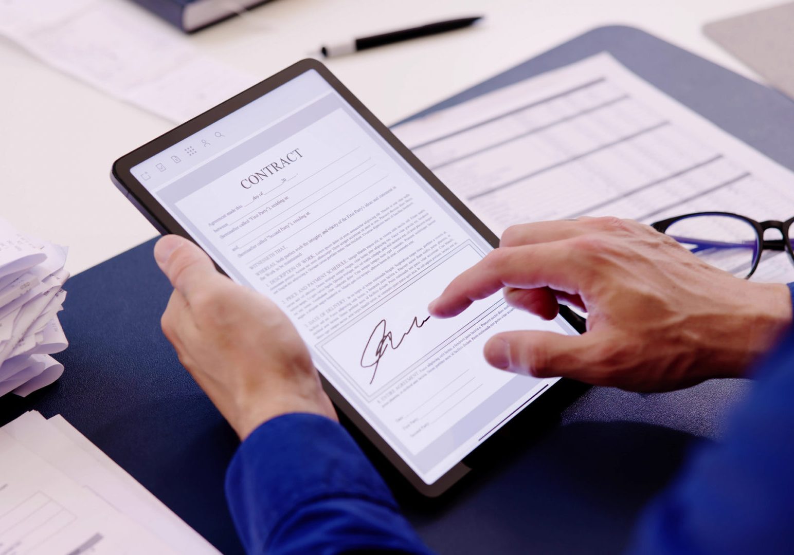 Person in blue shirt signing a digital contract document on a tablet, with glasses and paperwork on the desk nearby.