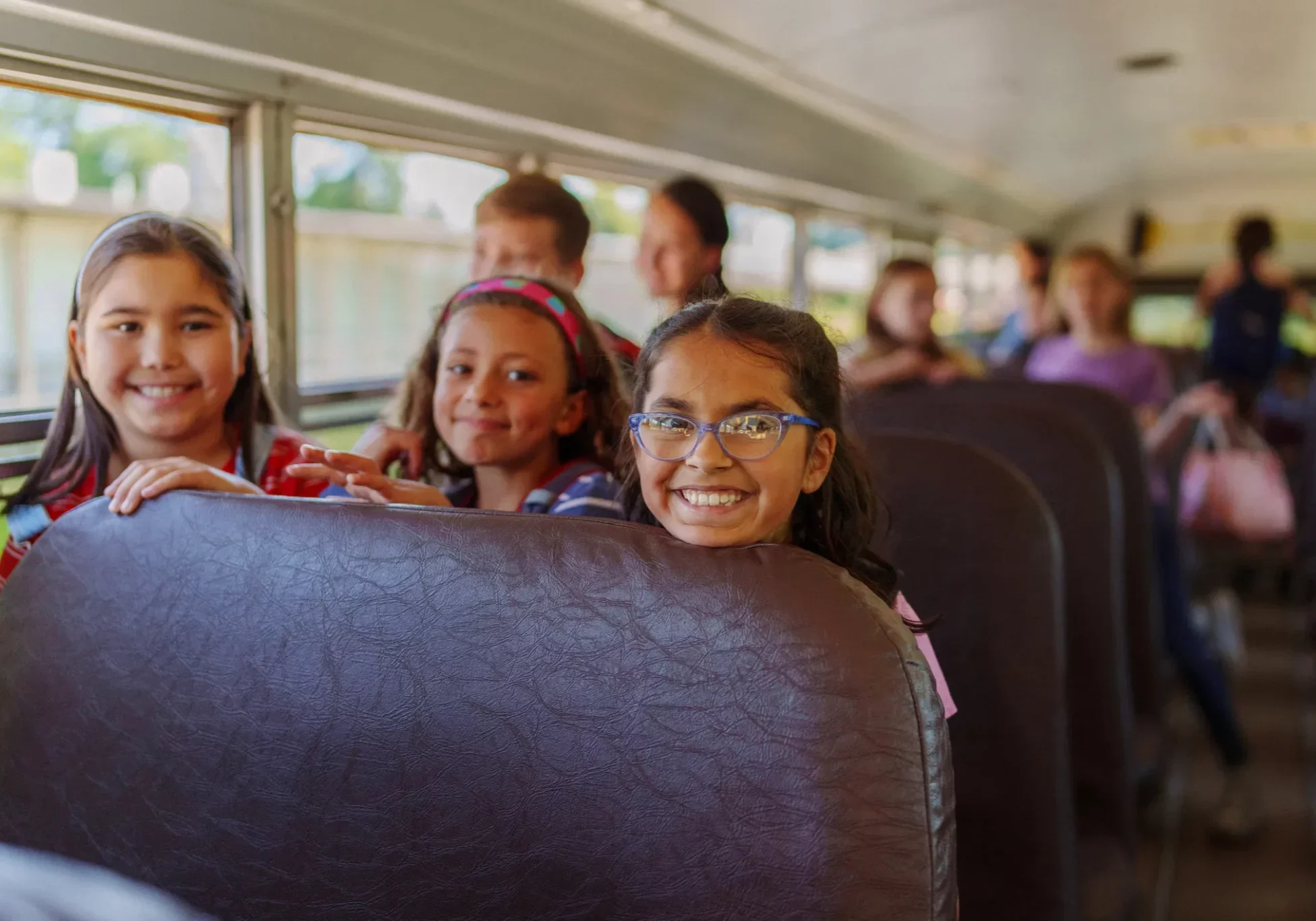 Three smiling elementary school girls riding a yellow school bus, with other students seated in the background.