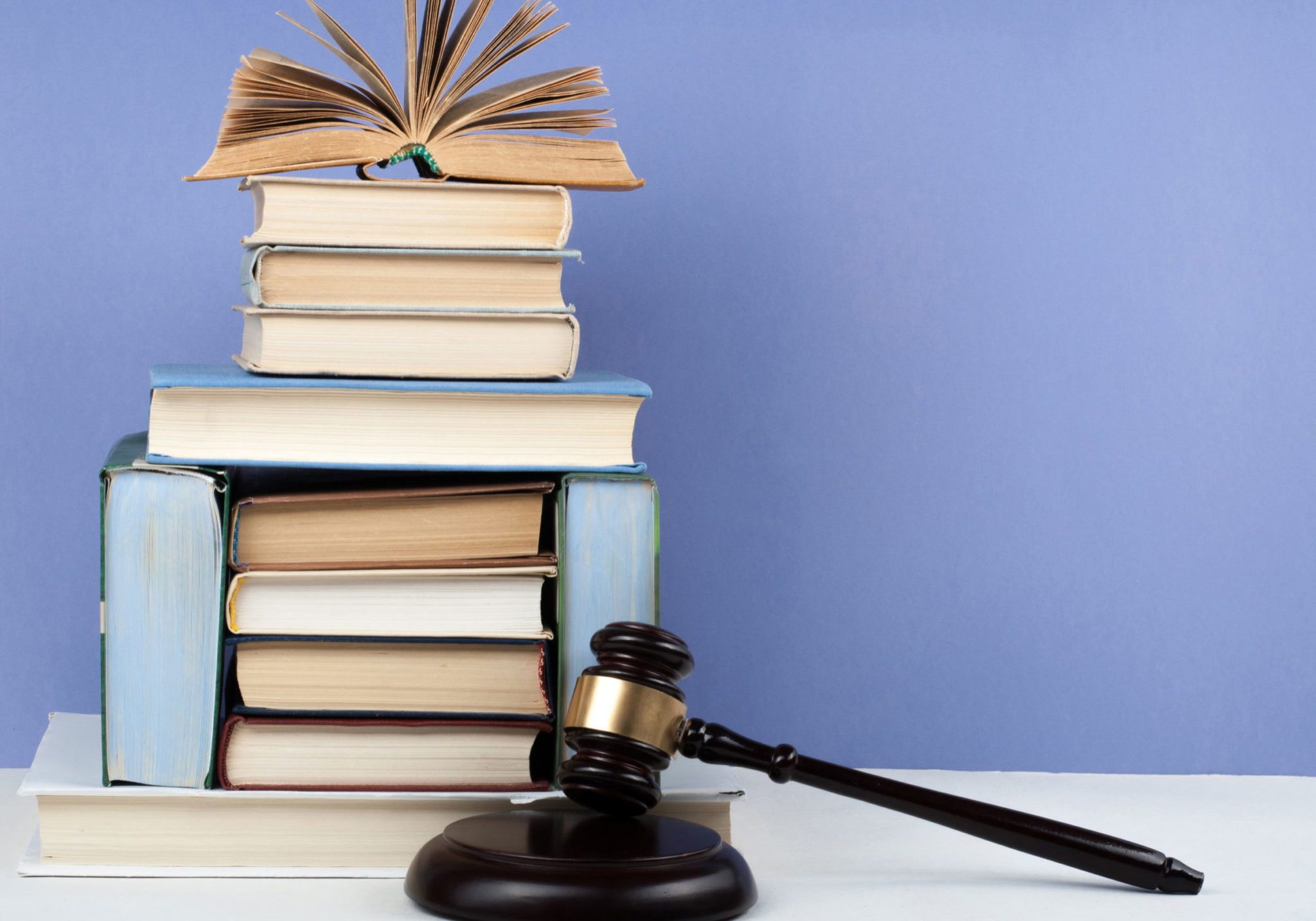 Wooden judge's gavel on a sound block beside a tall stack of law books, with one open book on top, against a blue background.