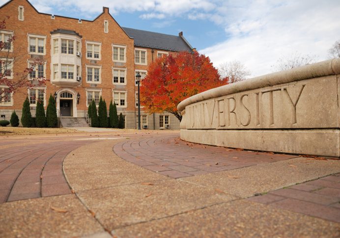 Stone monument engraved with "UNIVERSITY" on a brick campus plaza, with a red autumn tree and a three-story brick academic...