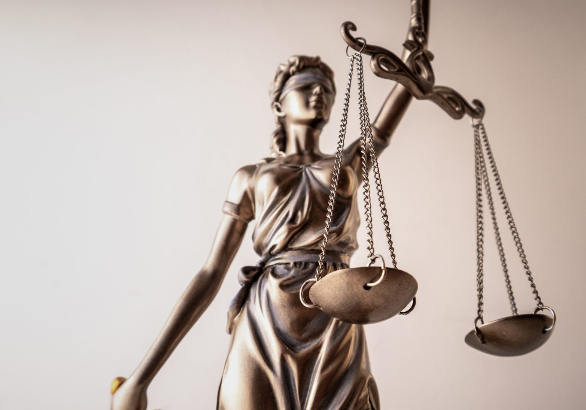 Bronze Lady Justice statue with blindfold, holding balanced scales in one hand and a sword in the other, against a light b...