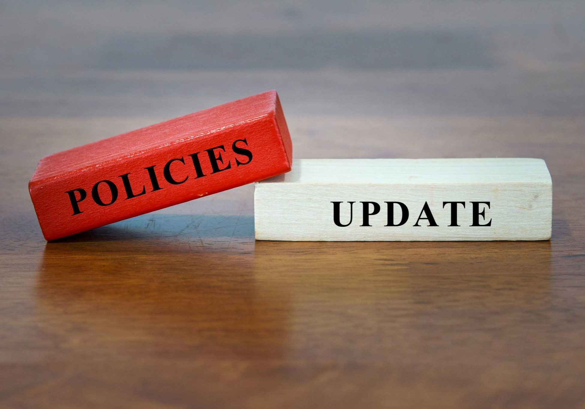 Two wooden blocks on a table, one red labeled POLICIES and one white labeled UPDATE, forming the phrase Policies Update