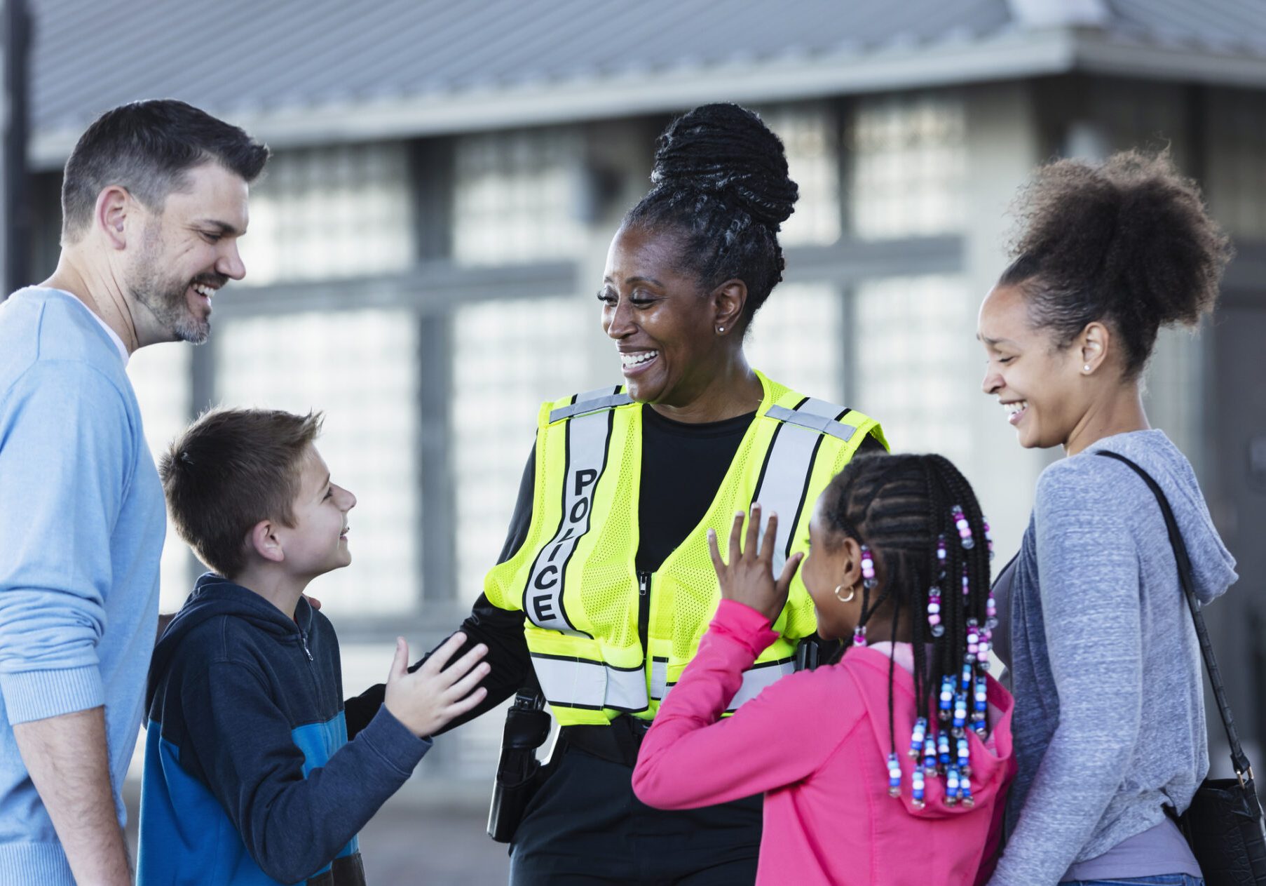 Smiling female police officer in yellow safety vest laughing with a family of four, including two children, outdoors near ...