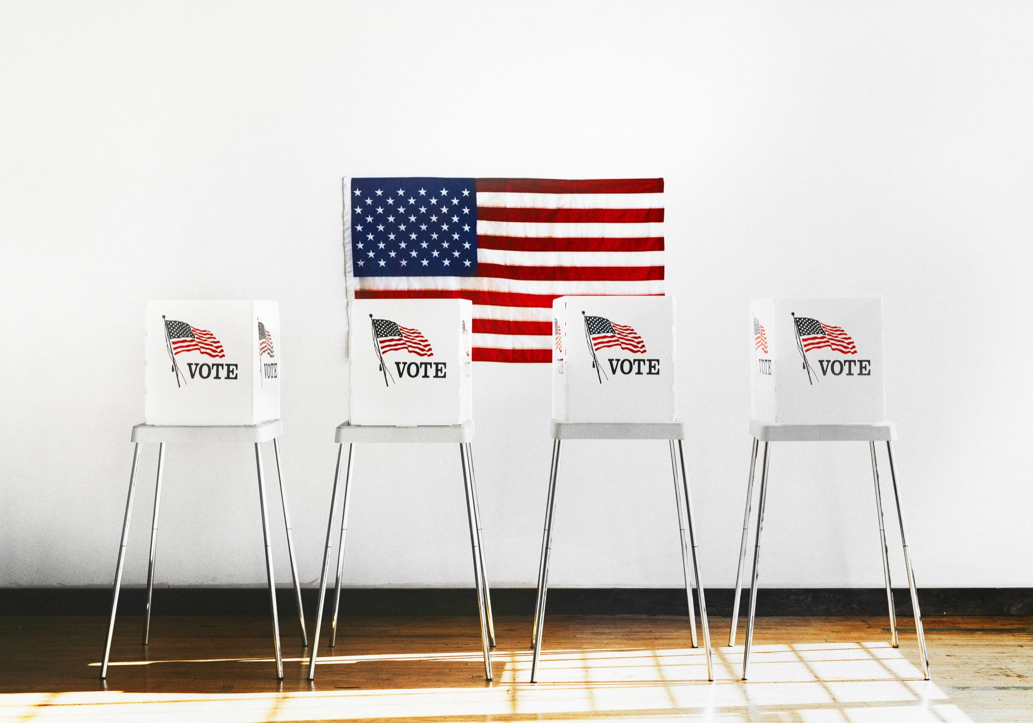 Four white voting booths labeled VOTE with American flag graphics, arranged in a row beneath a large US flag on a white wall