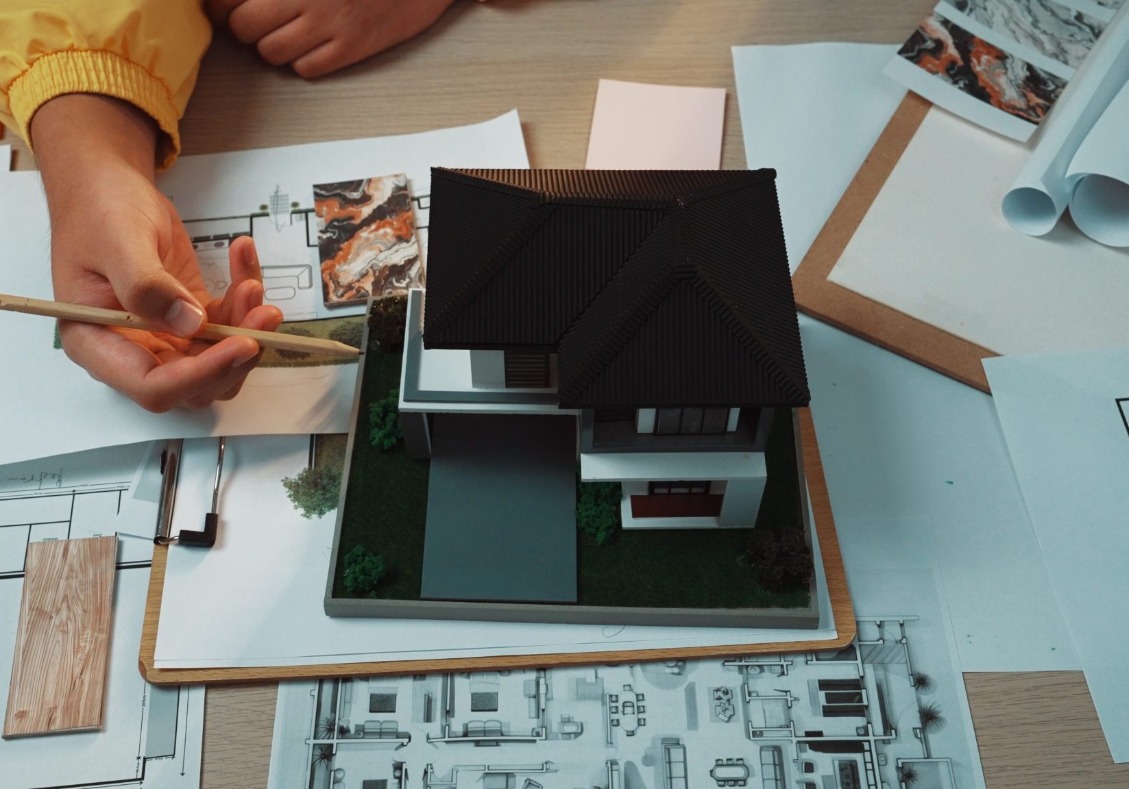 Architect's hands holding pencils over a detailed house scale model surrounded by blueprints, material samples, and rolled...
