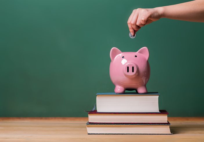 Hand dropping a coin into a pink piggy bank sitting on a stack of books in front of a green chalkboard
