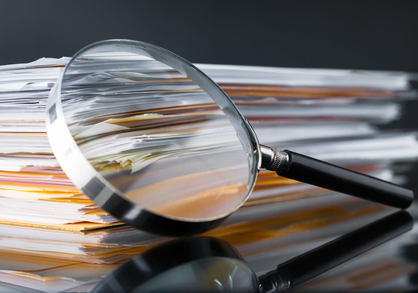Magnifying glass leaning against a large stack of white and yellow paper documents on a dark reflective surface
