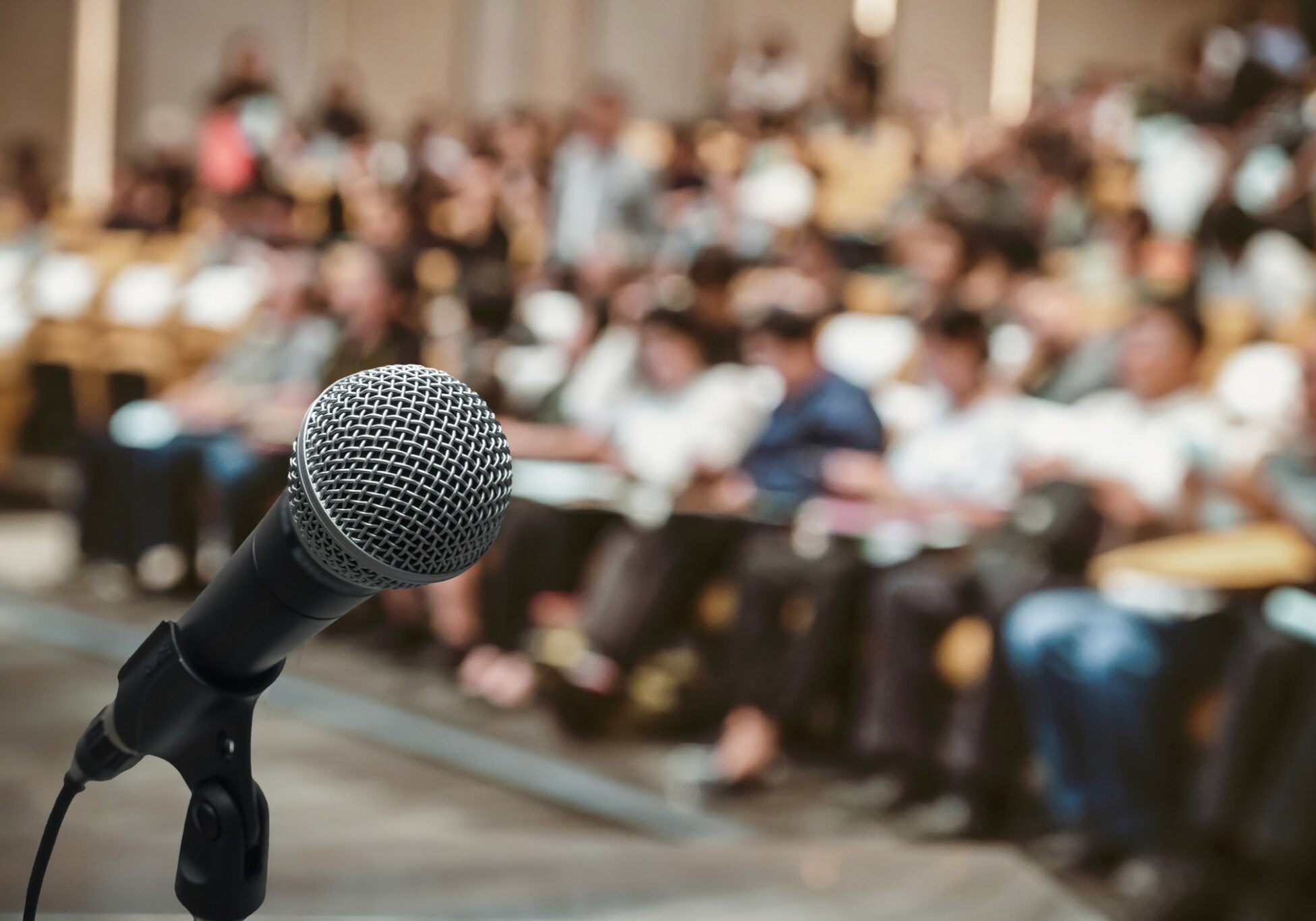 Black microphone on a stand in sharp focus with a large blurred audience seated in rows at a conference or event hall