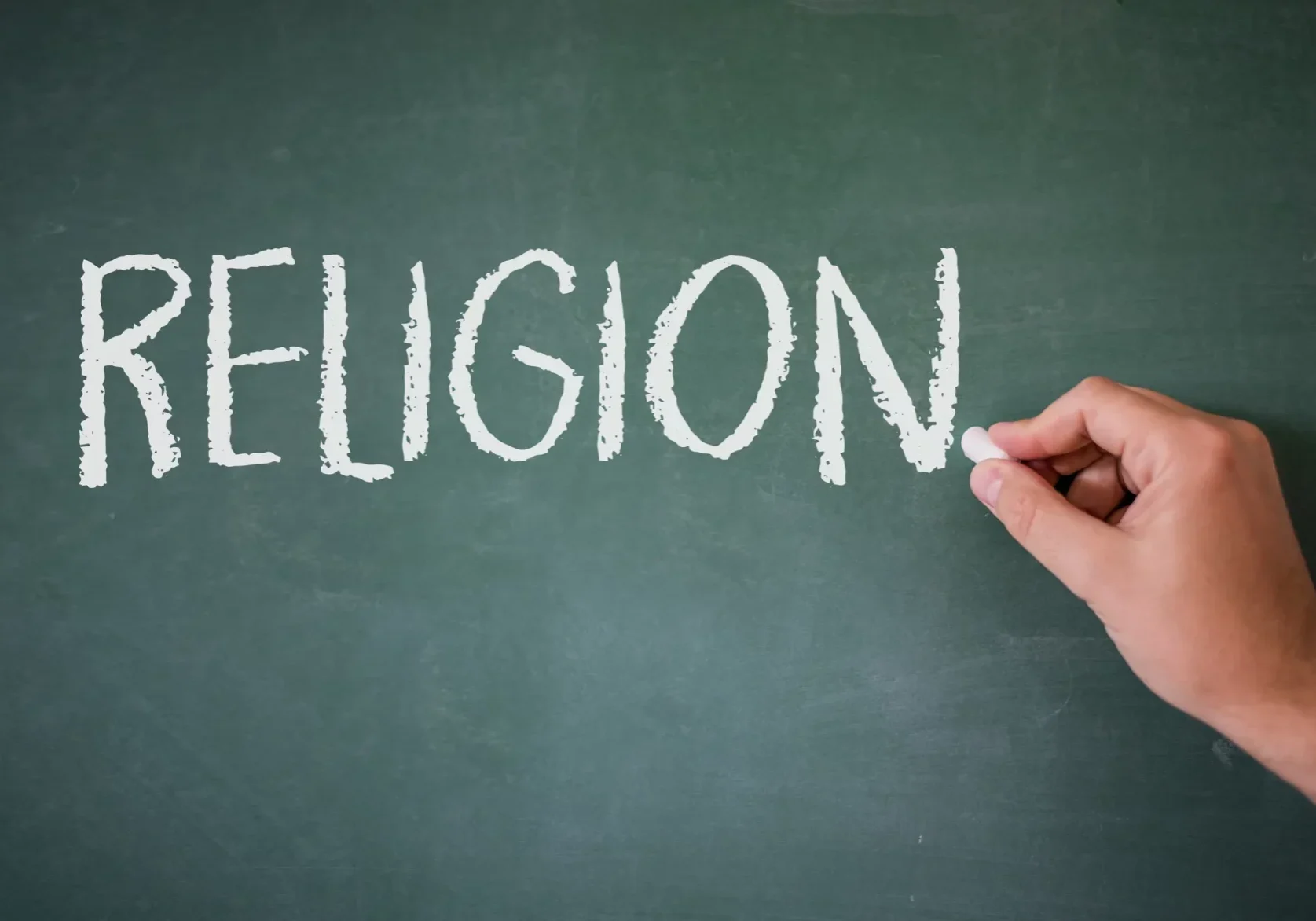 Hand writing the word RELIGION in white chalk on a green chalkboard