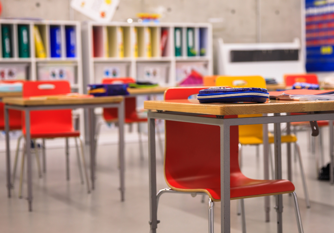 Empty elementary classroom with red and yellow chairs at wooden desks, colorful bookshelves, and Robbins Schwartz logo in ...
